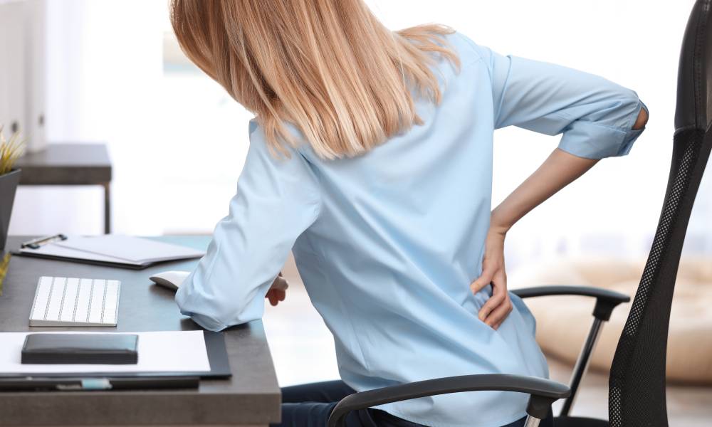 A woman at her desk reaching for her back in pain. She is working at her computer while sitting in a desk chair.